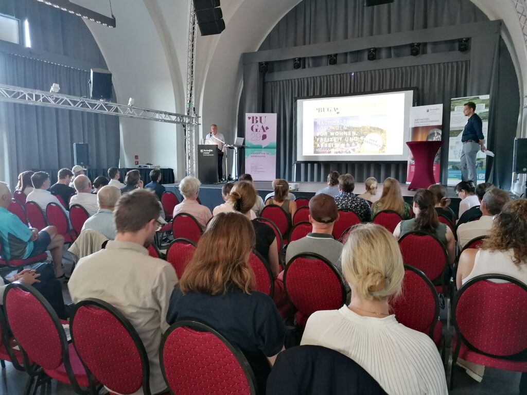 Viele Personen sitzen in einem Raum auf Stühlen und hören einem Vortrag zu.