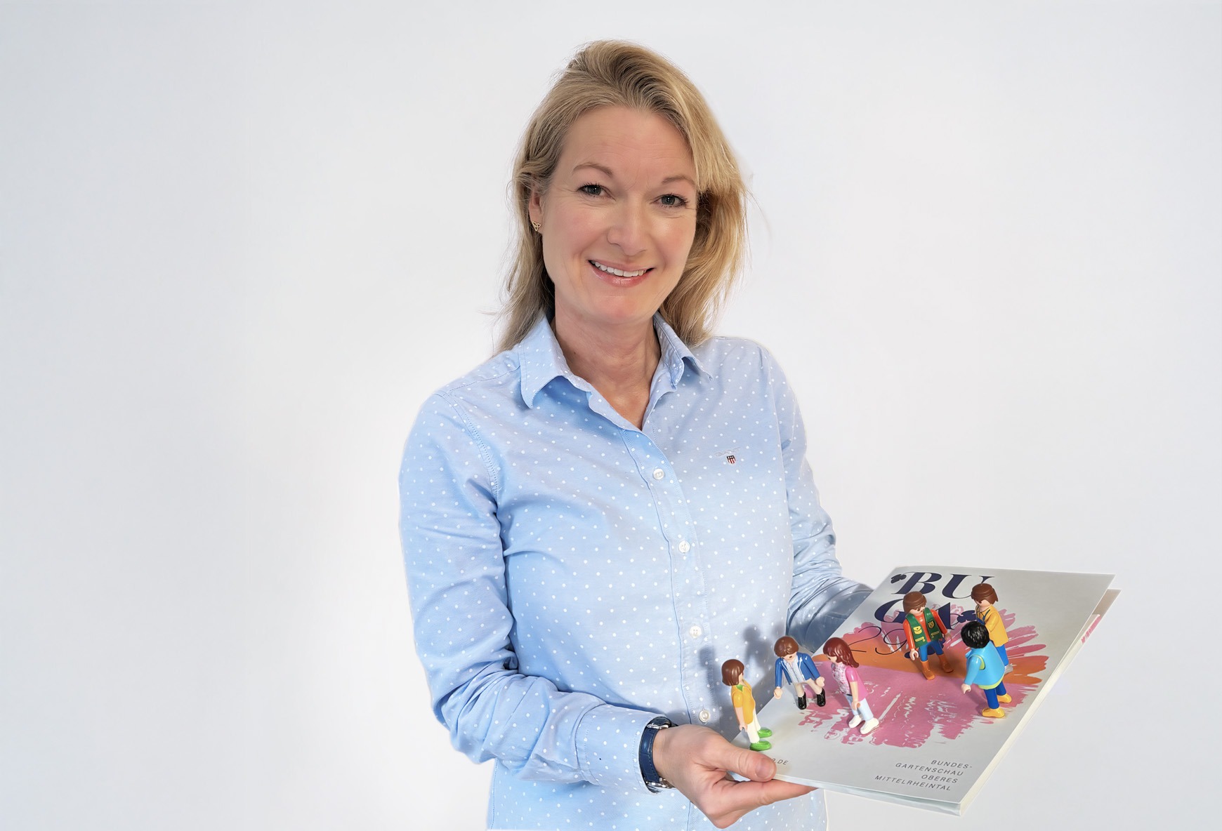 Angela Nass Porträt von Angela Nass, die eine BUGA-Broschüre hält, auf der Spielfiguren in verschiedenen Gesprächssituationen zu sehen sind.