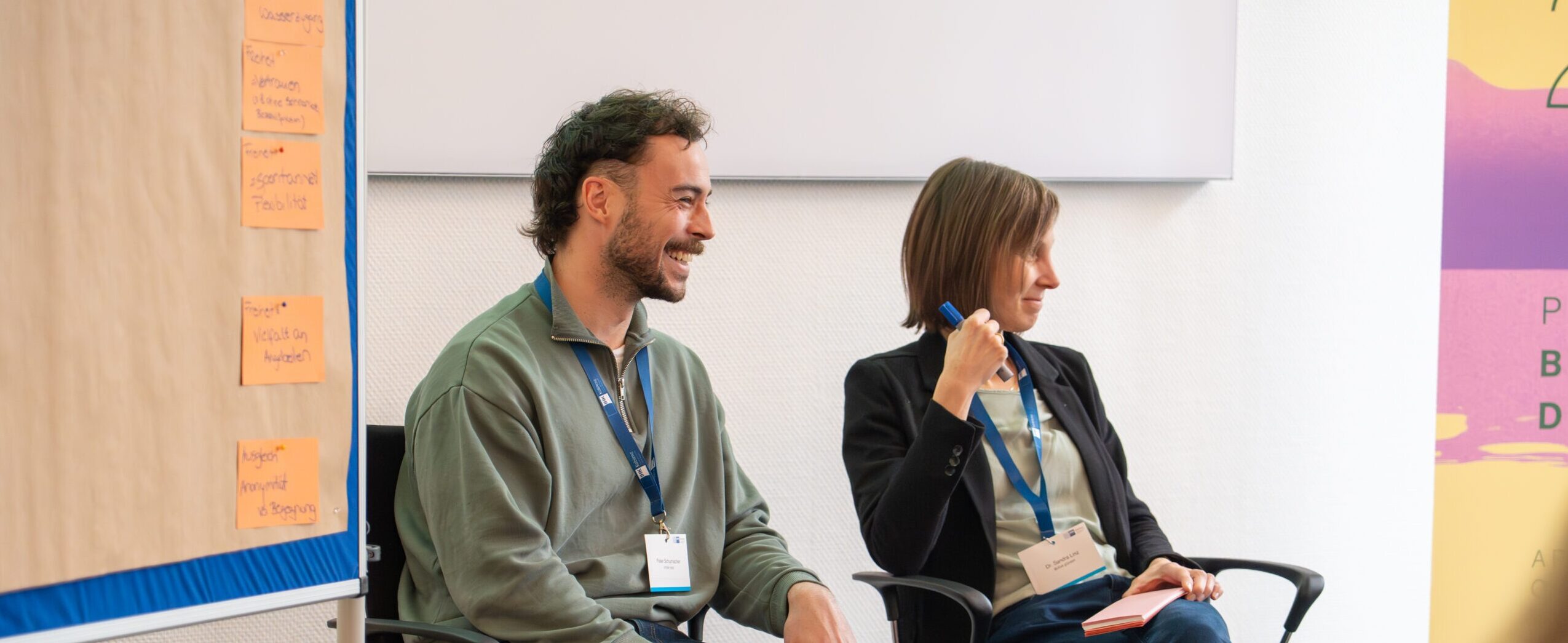 Zwei Personen sitzen in einem Workshop-Raum vor einer Pinnwand mit Moderationskarten und einem BUGA-Roll-up.
