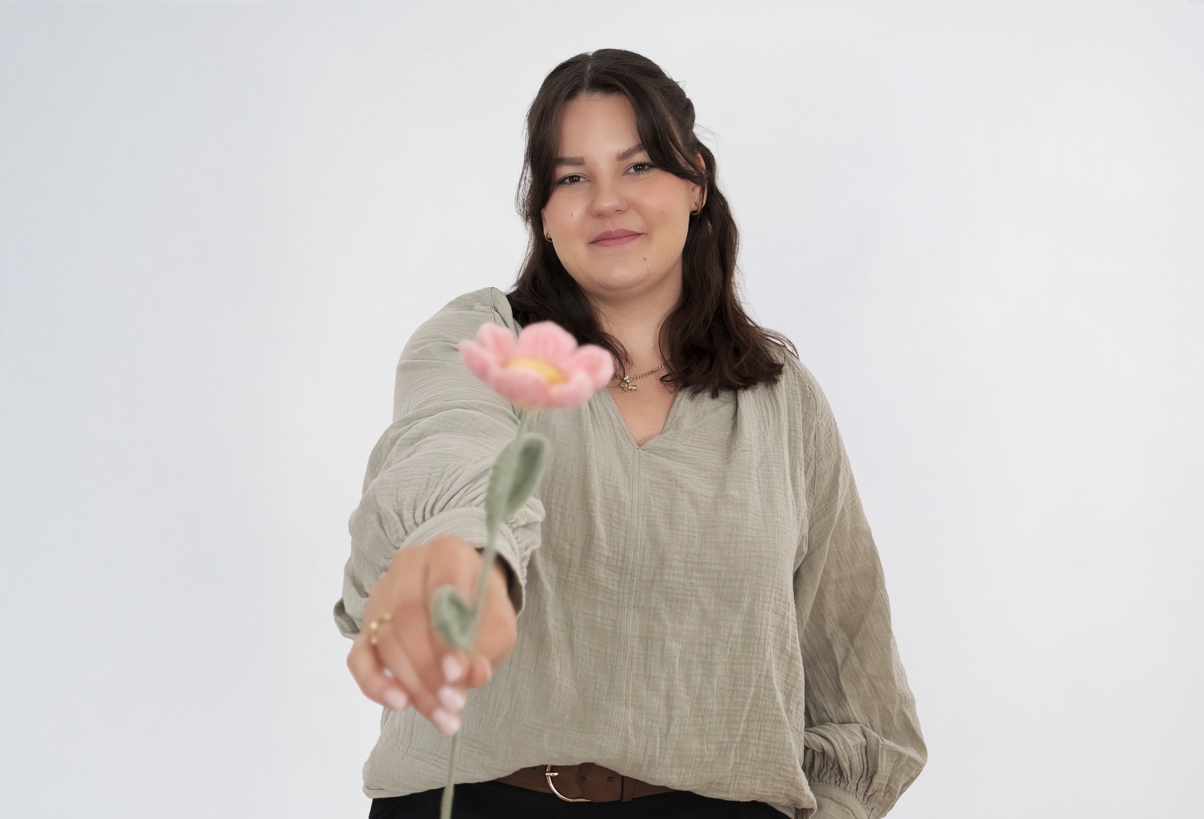 Eine Frau mit braunen Haaren und einer grünen Bluse lächelt und hält eine Häckelblume in der Hand.