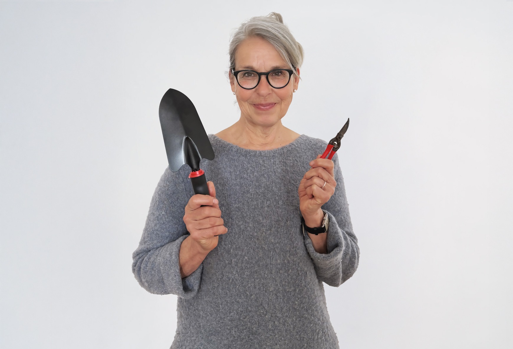 Eine blonde Frau in einem grauen Pullover hält eine Gartenschaufel und Gartenschere in den Händen und lächelt.