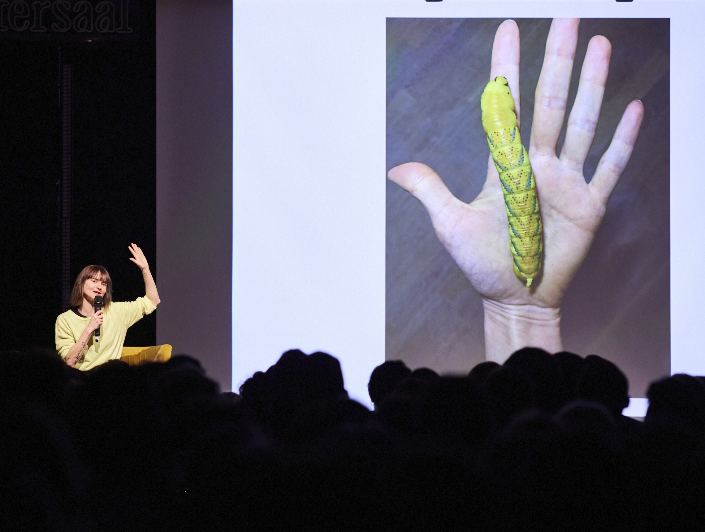 Eine Frau spricht mit Mikrofon auf einer Bühne vor Publikum; auf der Leinwand hinter ihr ist eine große grün-gelbe Raupe auf einer Hand zu sehen.