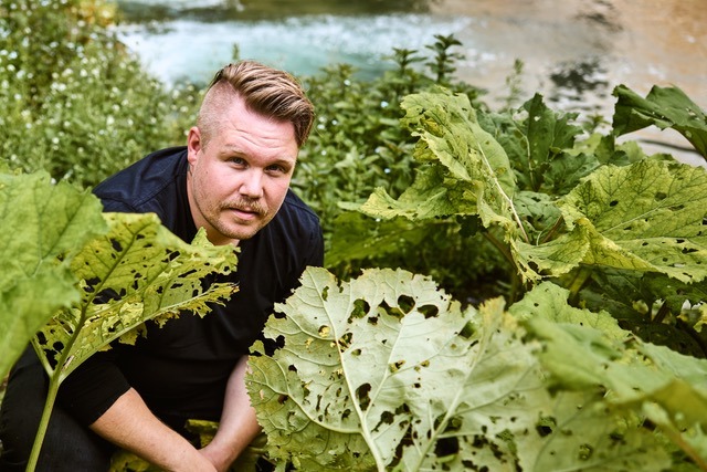 Thomas-Hörren_Credits_Maximilian Kamps-Agentur Blumberg Ein Mann hockt zwischen großen grünen Blättern in einem Gartenbeet, im Hintergrund fließt Wasser