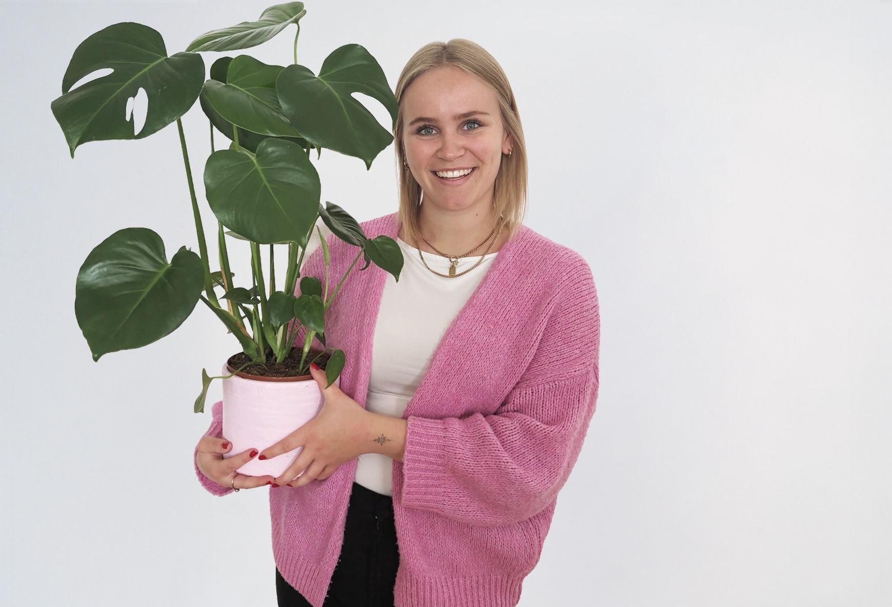 Katharina_2 Eine Frau mit blonden Haaren und einer rosa Strickjacke lächelt und hält eine Pflanze in den Händen.