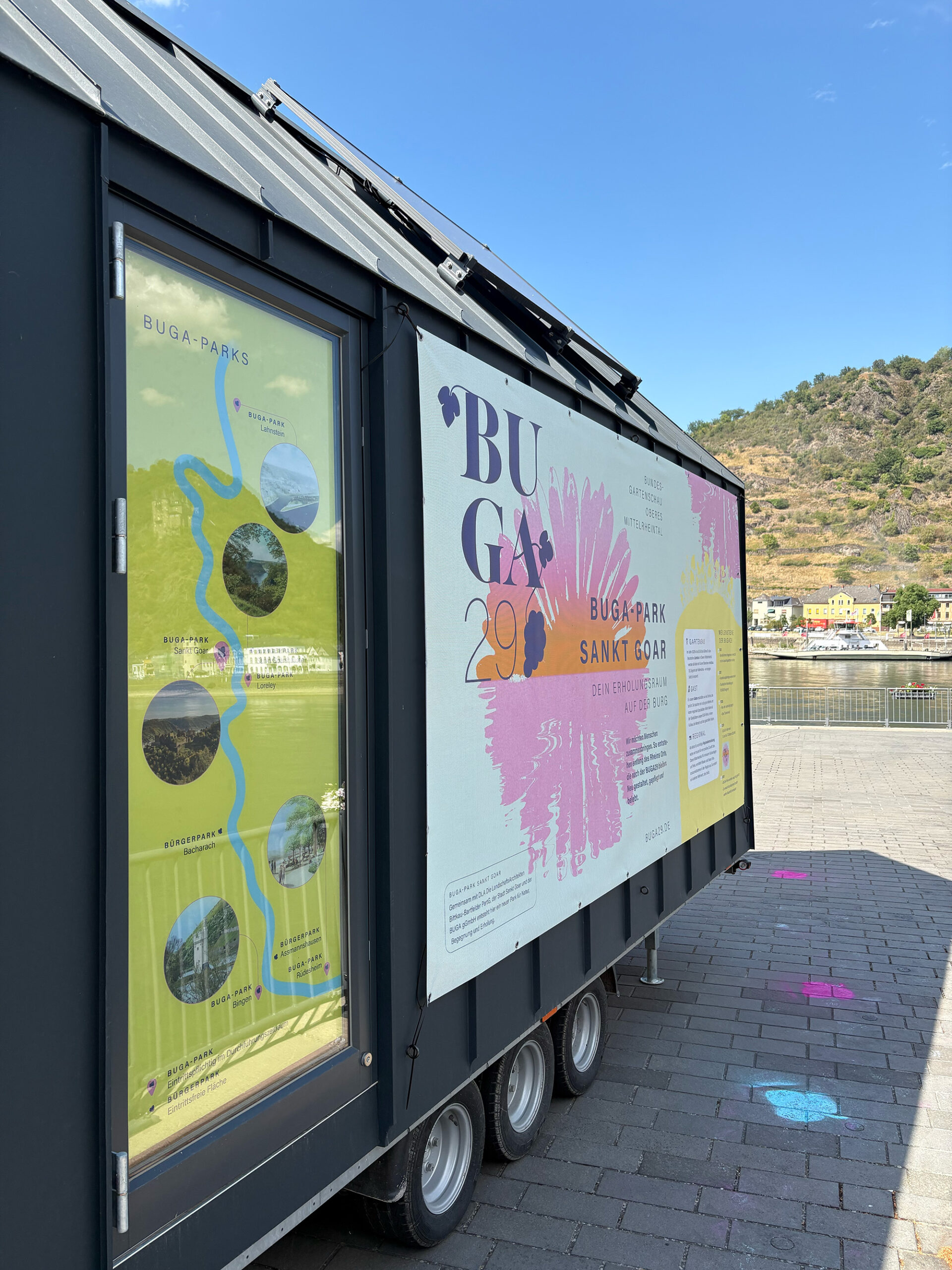 Seitenansicht des BUGA29-Tiny-Houses am Rhein mit einer Rheinkarte aller BUGA-Parks im Fenster und dem großen Banner „BUGA-Park Sankt Goar – Dein Erholungsraum auf der Burg".