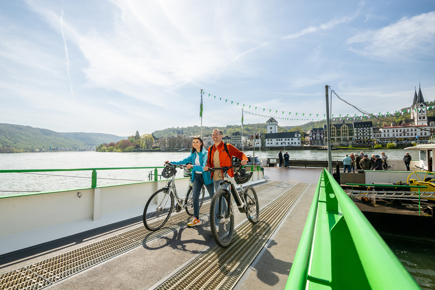 Radfahrer auf einer Rheinfähre Zwei Radelnde mit Helmen machen Pause auf einer Steinmauer am Rhein und schauen auf den Fluss, dahinter ein bewaldeter Hang mit Burg.