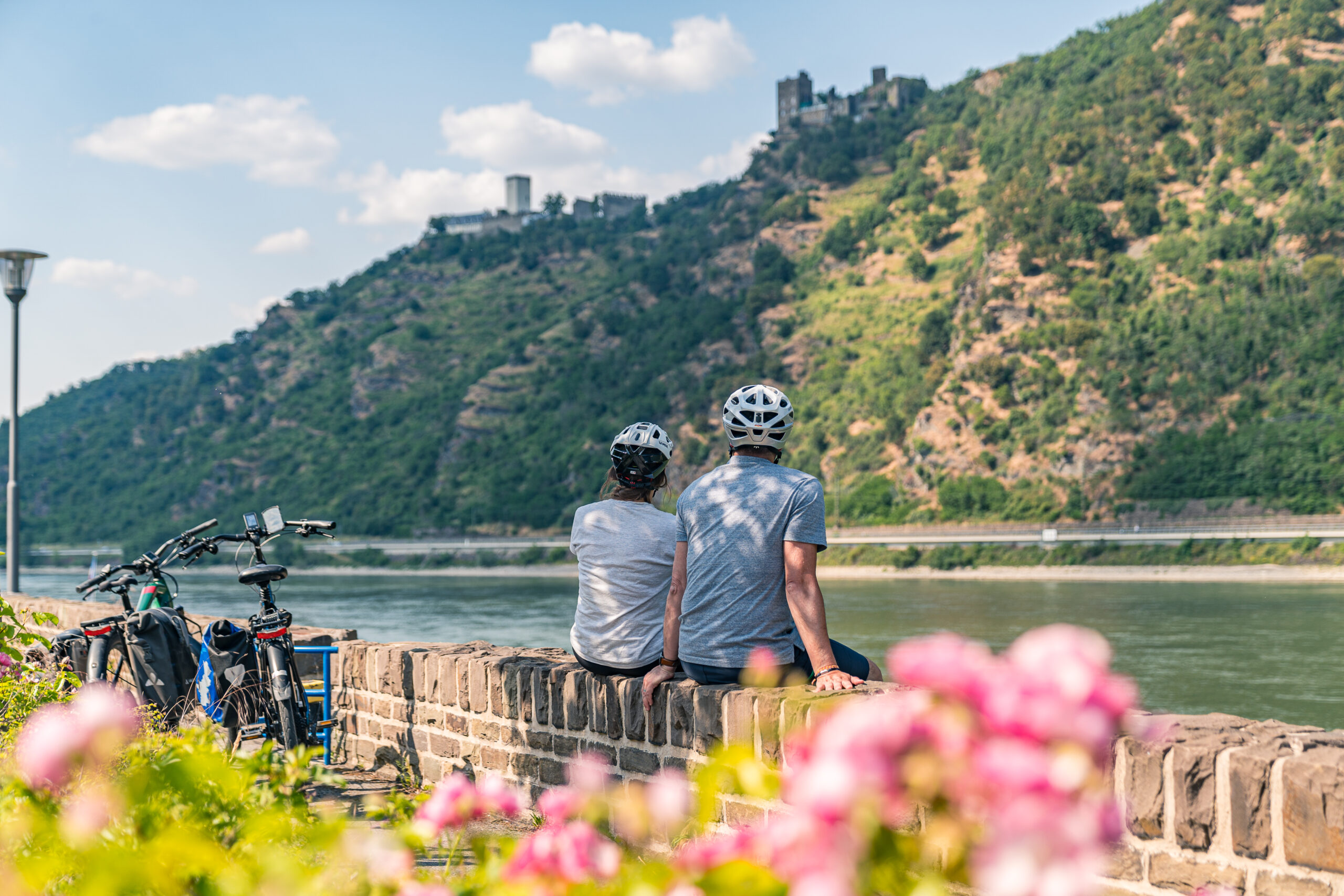Zwei Radelnde mit Helmen machen Pause auf einer Steinmauer am Rhein und schauen auf den Fluss, dahinter ein bewaldeter Hang mit Burg.