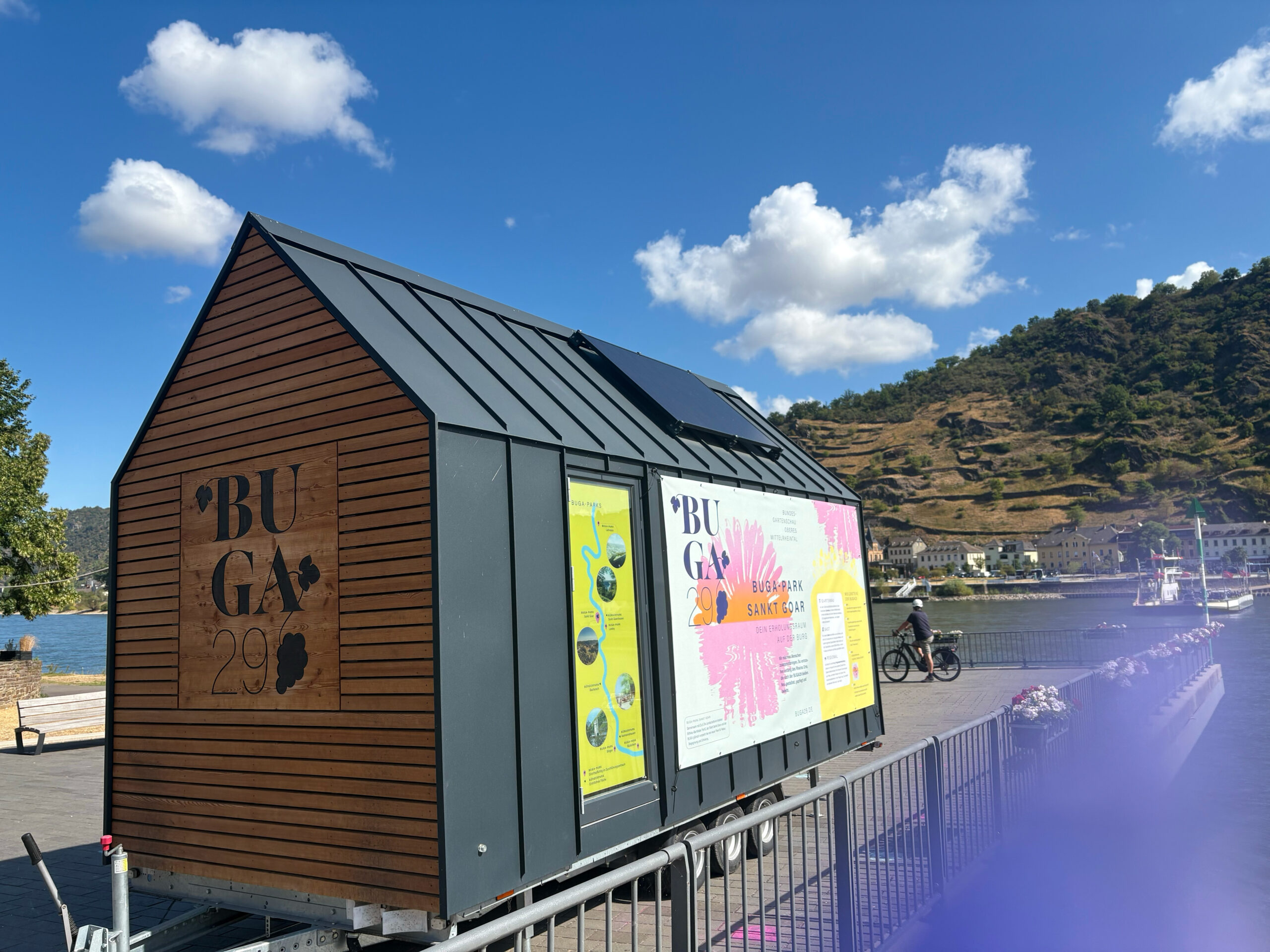 Seitenansicht des BUGA-Tiny-Houses am Rhein mit einer Rheinkarte aller BUGA-Parks im Fenster und dem großen Banner „BUGA-Park Sankt Goar – Dein Erholungsraum auf der Burg".